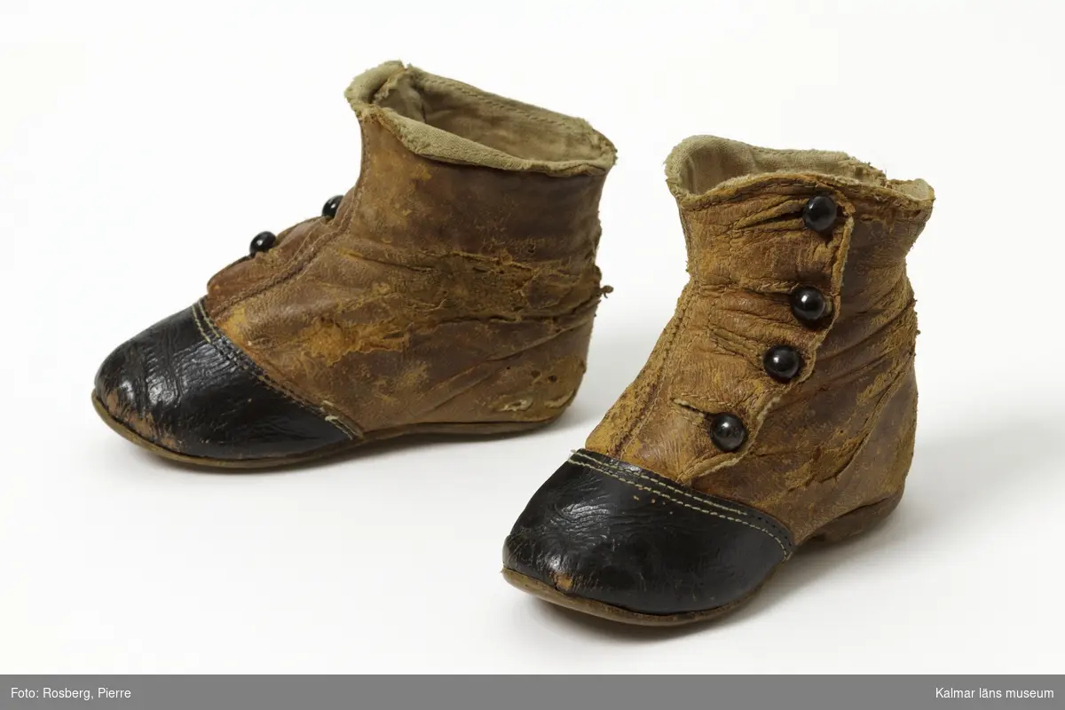 KLM 31675:1-2. Skor, barnkängor, ett par, av skinn, låg läderklack, lädersula och fodrad med beige bomullstyg. Skinn, brunt vid fot, svart tåhätta. Knappknäppning. Knäpps med fyra svarta knappar och knapphål. Klacken saknas på höger sko. Sliten.