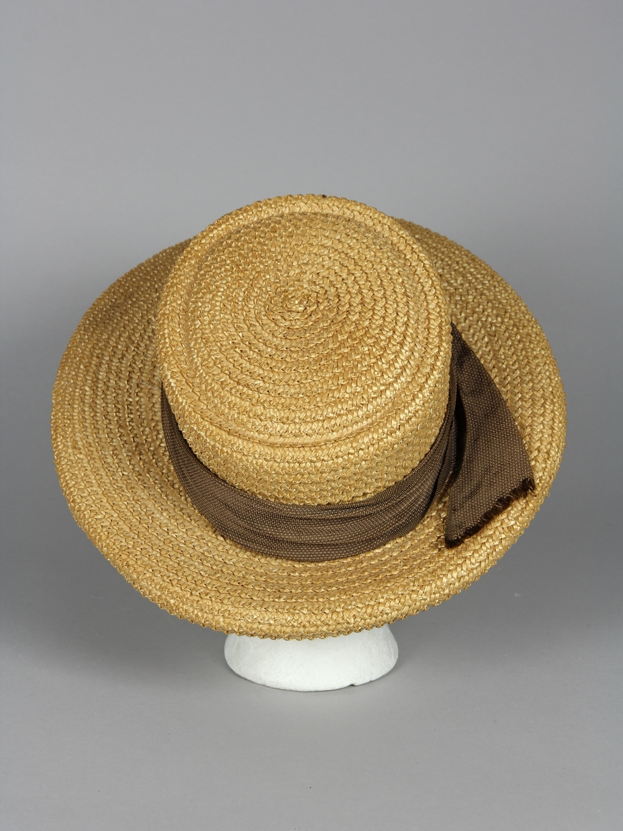 Stråhatt av flätat ståband. Med ett asymetriskt monterat hattband i brunt med tunna ljusa linjer. Hattens brätte är bredare framtill än baktill. Brättet har en svagt klockad form. Kullen är oval och med flat avslutning.