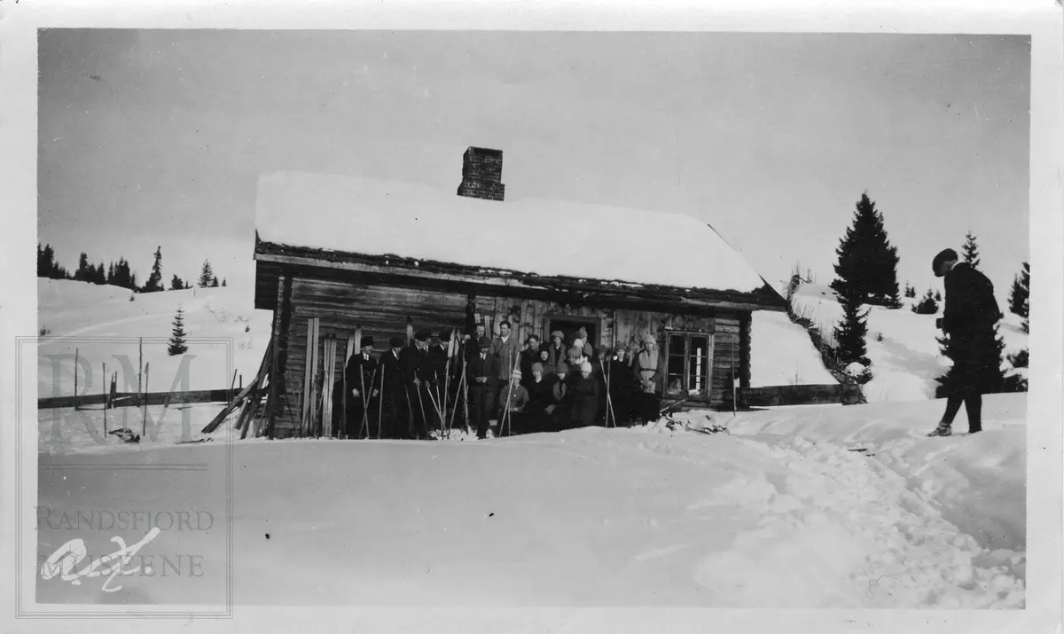En gruppe ungdom og voksne med ski samlet utenfor ei tømmerstue eller hytte. Fotograf til høyre.