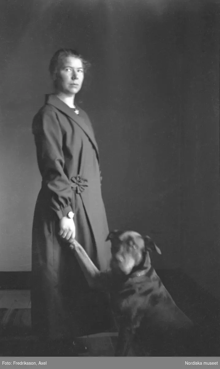Fogelstad. Emmy Karlsson avporträtterad stående i helfigur. Bredvid henne sitter Elisabeth Tamms hund Pojken och hon håller hans högra tass i sin högra hand .