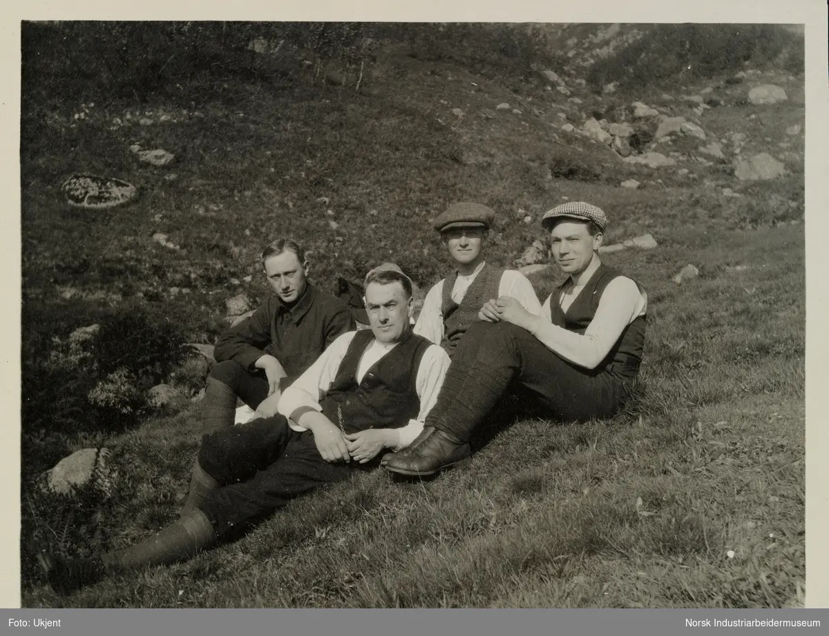Tre menn sitter og en mann ligger på gress i naturen