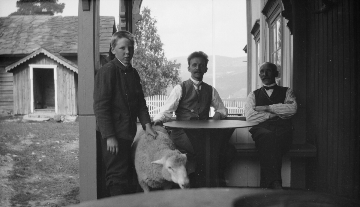 To mann, en gutt og en sau ved bord på verandaen, antakelig på Linjordet i Østre Gausdal