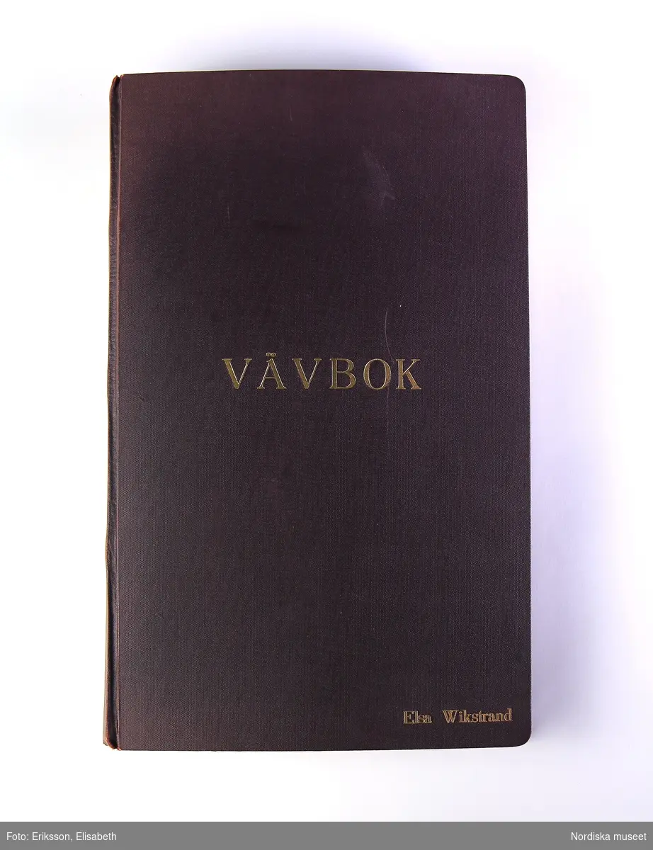 Vävbok. Svart pärm med tryck i guldfärg på framsidan "VÄVBOK" / "ELSA WIKSTRAND". Boken är handskriven med text och teckningar av vävstolar och vävnotor. Inklistrade tygprover.