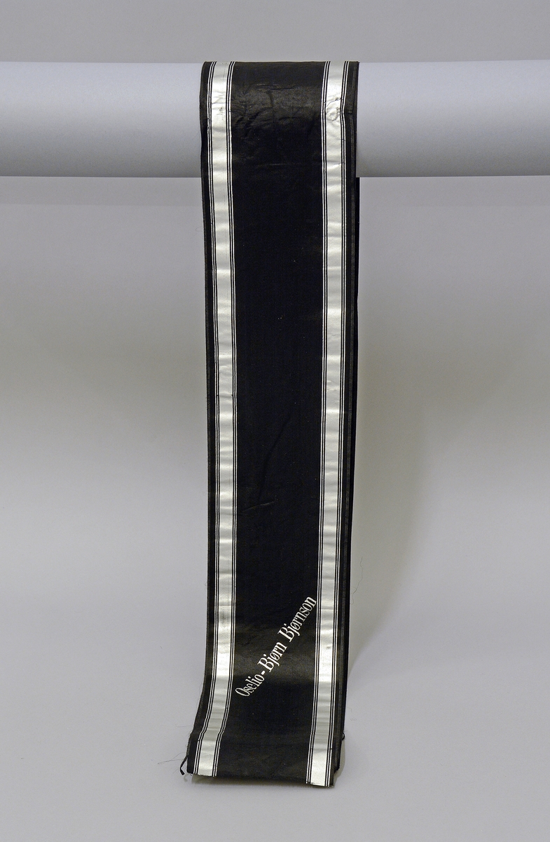 Båndet er sort med sølvstriper og sølvskrift.