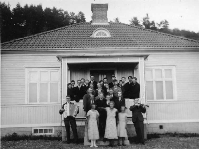 Okänd grupp av män och kvinnor står på farstun till ett hus i Uppgränna. Nedanför trappan står till vänster en man med dragspel och till höger en man med fiol.