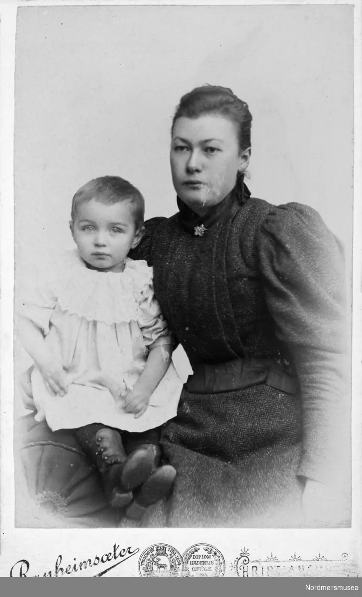 Portrettfoto av en mor med et barn på fanget. Trolig fra Kristiansund, siden det er der han er fotografert. Det er Ole Ranheimsæter som er fotograf. Fra Nordmøre museums fotosamlinger.