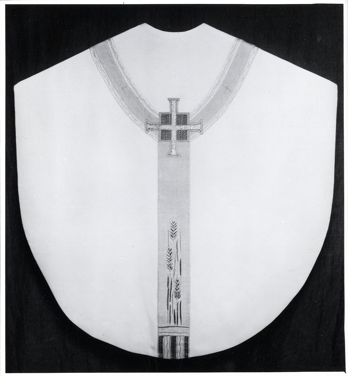 Foto (svart/vitt) av en ljus mässhake (baksidan) med något mörkare, broderat korsparti, prytt med bl a sädesax och liksidiga kors.

Inskrivet i huvudbok 1983.