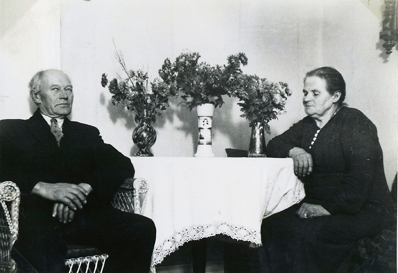 Johan Karlsson (1866 - 1947) och hustrun Augusta 1869 - 1957) sitter i sitt hem bredvid ett bord där det står tre vaser med blombuketter, Hallen 1:4 "Johans", okänt årtal.