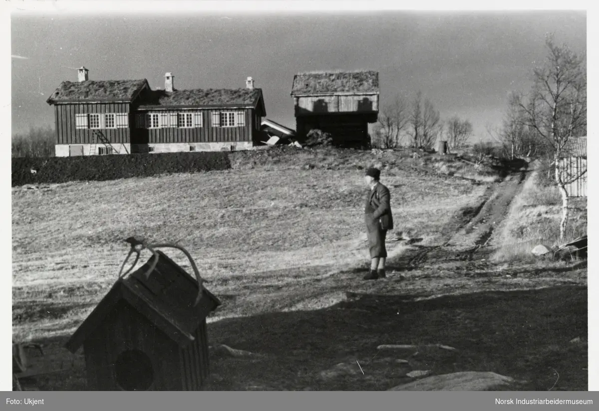Mann med hatt står ved gårdsvei opp til Sundet, Møsstrond. Fremst sees et hundehus med gevir plassert på taket.