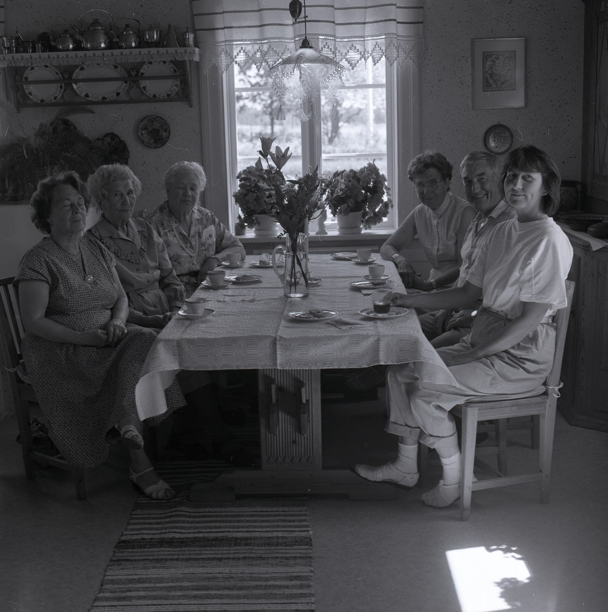 Vid ett vackert dukat bord sitter Adéle med några kvinnor och en man på stolarna, på bordet ligger en vacker duk och det står fina blommor i en vas där och kaffekoppar med fat. En matta ligger det på golvet, i fönstert är det en gardin, blommor och en lampa ovanför bordet. På väggen hänger en tallrikshylla med kopparföremål, tallrikar och tavlor. Från Adéles 70-årsdag 16 juni 1988.