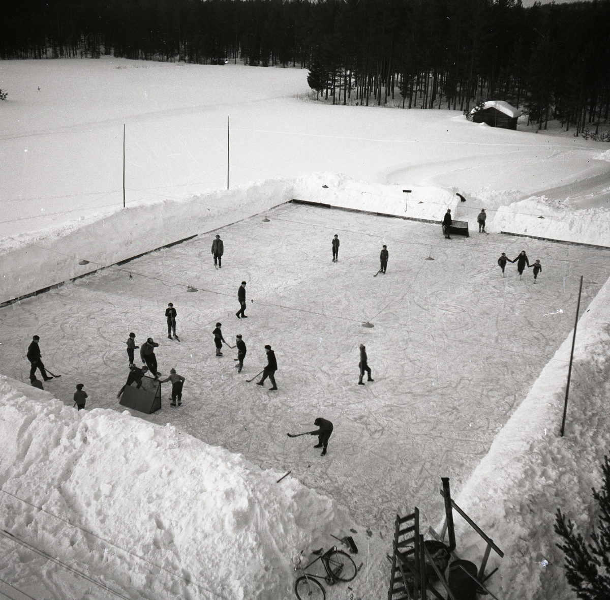 På isplan åks det skridskor och spelas ishockey av en grupp människor. Det finns höga snövallar på isplanens kanter, hockeyklubbor, målburar, cykel, stege, skog och en lite byggnad. Det finns även belysning ovanför isplan. Ishockey Glösboplanen 27 januari 1959.