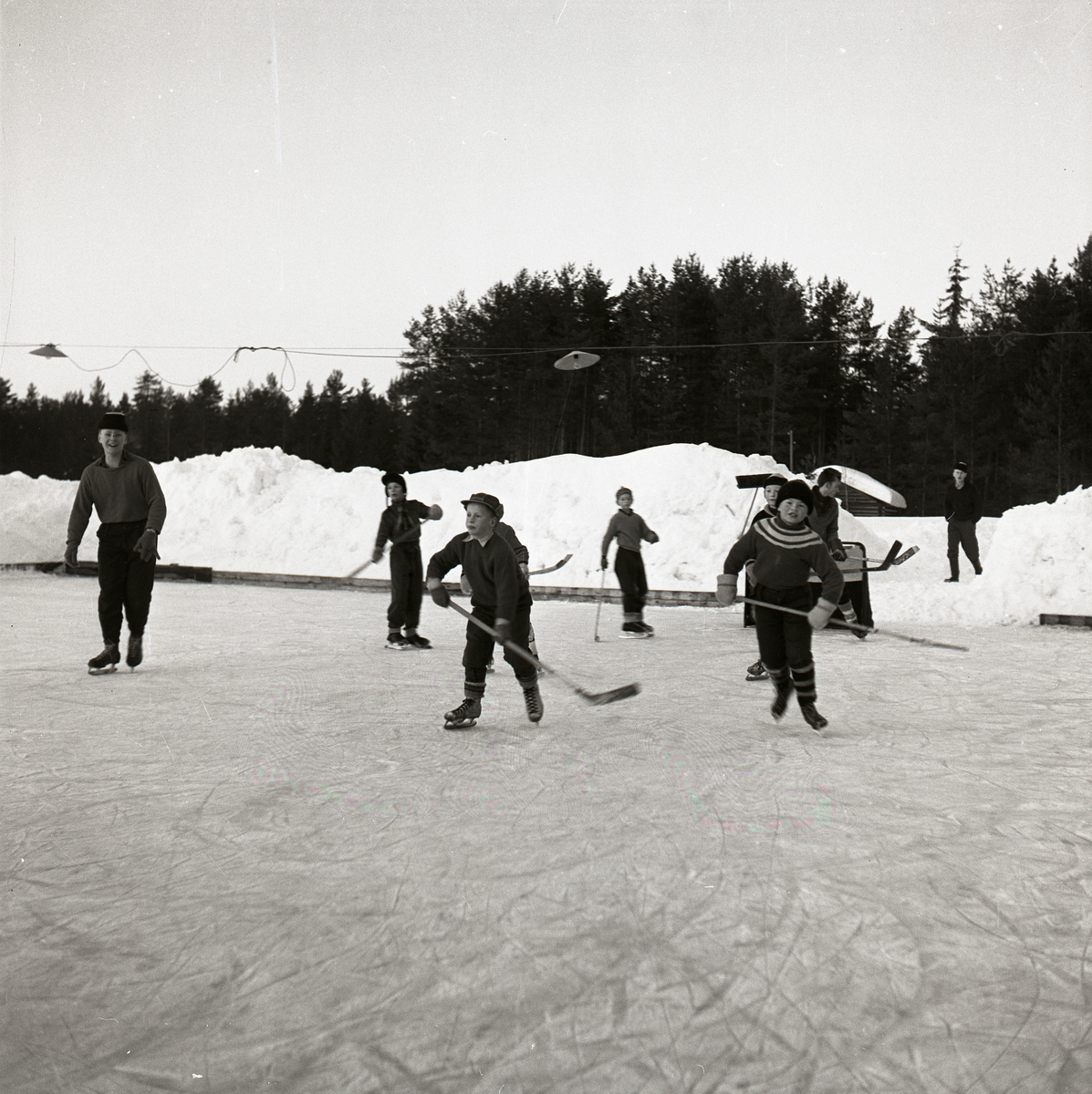 Några ishockeyspelande pojkar med hockeyklubbor och skridskor. Det är en isplan med sarg, belysning, snöhögar, skog, himmel och en liten byggnad. Ishockey Glösboplanen 27 januari 1959.