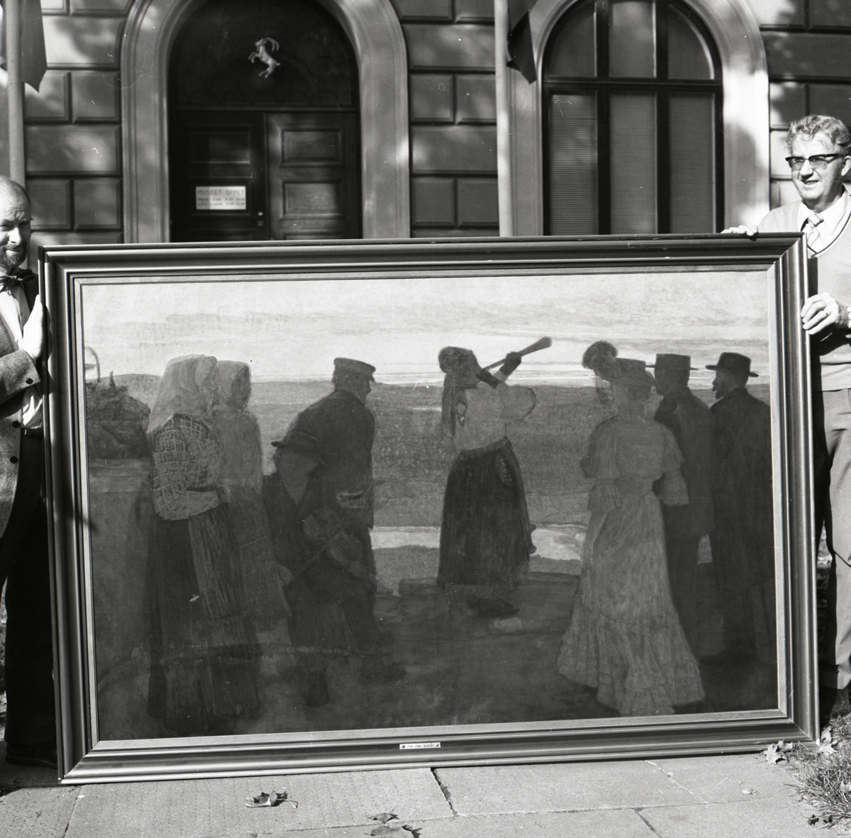 Två män håller upp tavlan "På blacksås" målad av John Sten. Framför entrén till Hälsinglands museum, 1974-1975.