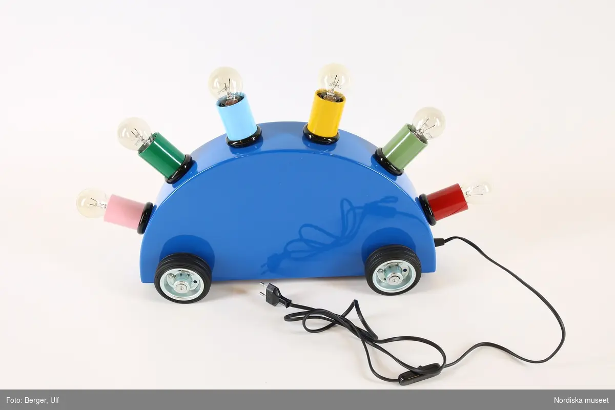 a-g)
a) Bordslampa, elektrisk, nytillverkad, i form av enkel leksaksbil uppdragen i skala. Chassi i blåmålad plåt: konvex rygg med sex glödlampshållare i rött, ljusgrönt, gult, ljusblått, mörkgrönt och rosa, alla med iskruvade b-g) klara glödlampor. Fyra svarta gummihjul. Sladd längst fram på ena kortsidan, påminnande om dragsnöre på leksaksbil.

Kring dragsnöret två klisteretiketter, text se nedan.
/Maria Maxén 2017-06-28