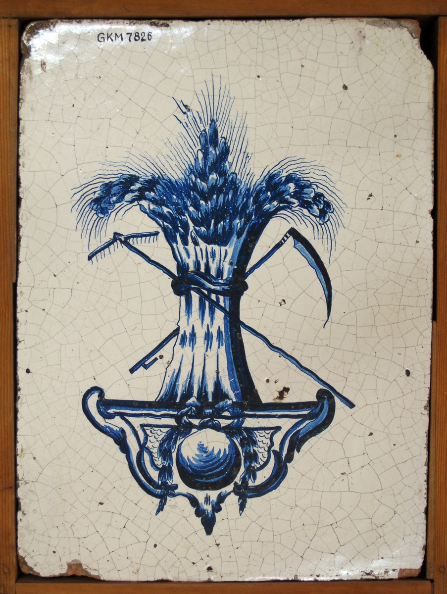 Acc. kat: Kakelplatta, vit glasyr, dekor i blått: kärve med räfsa och lie i heraldisk placering över rokokovoluter.
