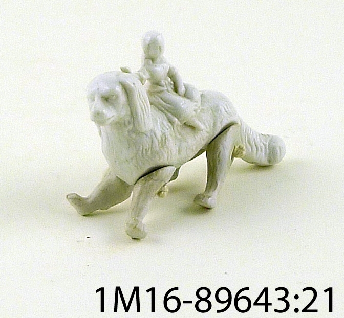 Figurin, vit, hund av porslin med ledade ben, på ryggen sitter en flicka eller docka föreställande en flicka.
