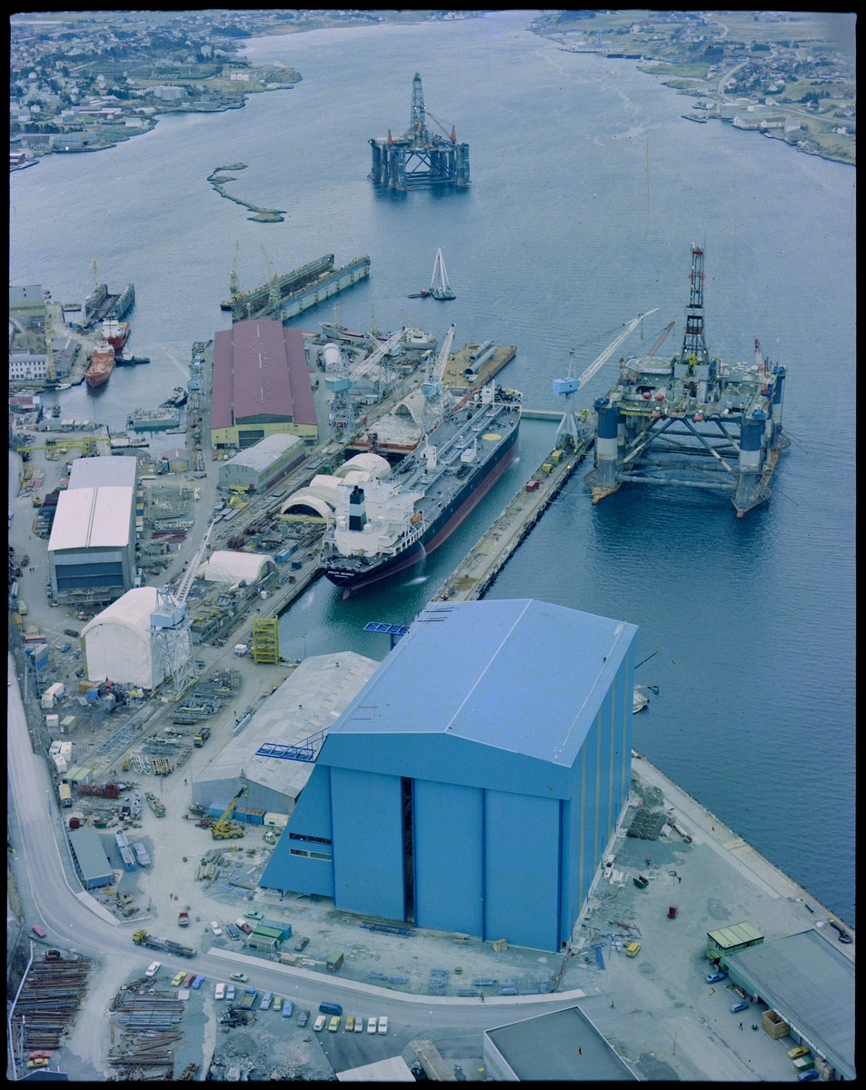 Flyfoto av Haugesund Mekaniske Verksted med omegn. Et stort skip ligger i tørrdokken, riggen "Ocean Liberator" ligger rett utenfor og en rigg ligger ved Storasundskjer. Nordsjøhallen er ferdig og tatt i bruk.