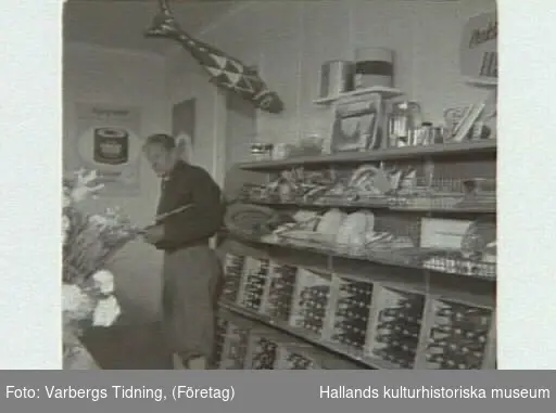 Affärsinteriör med staplade drickabackar i trä och en man i "äppelknyckarbyxor". "Kooperativa öppnar ny affär på Getterön". Artikel i samband med bilden publicerad i Varbergs Tidning 1955-06-15.