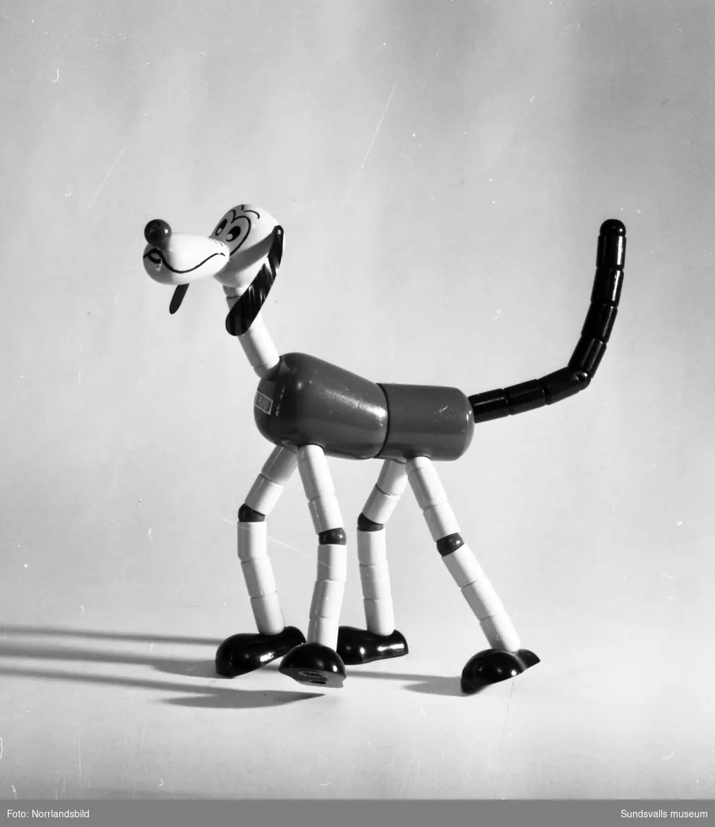 Olika leksaker fotograferade för varuhuset Dahlmans julannonsering 1959. Bland annat en klocka med kulram, docka, bilar i trä och plåt samt en hund i trä.