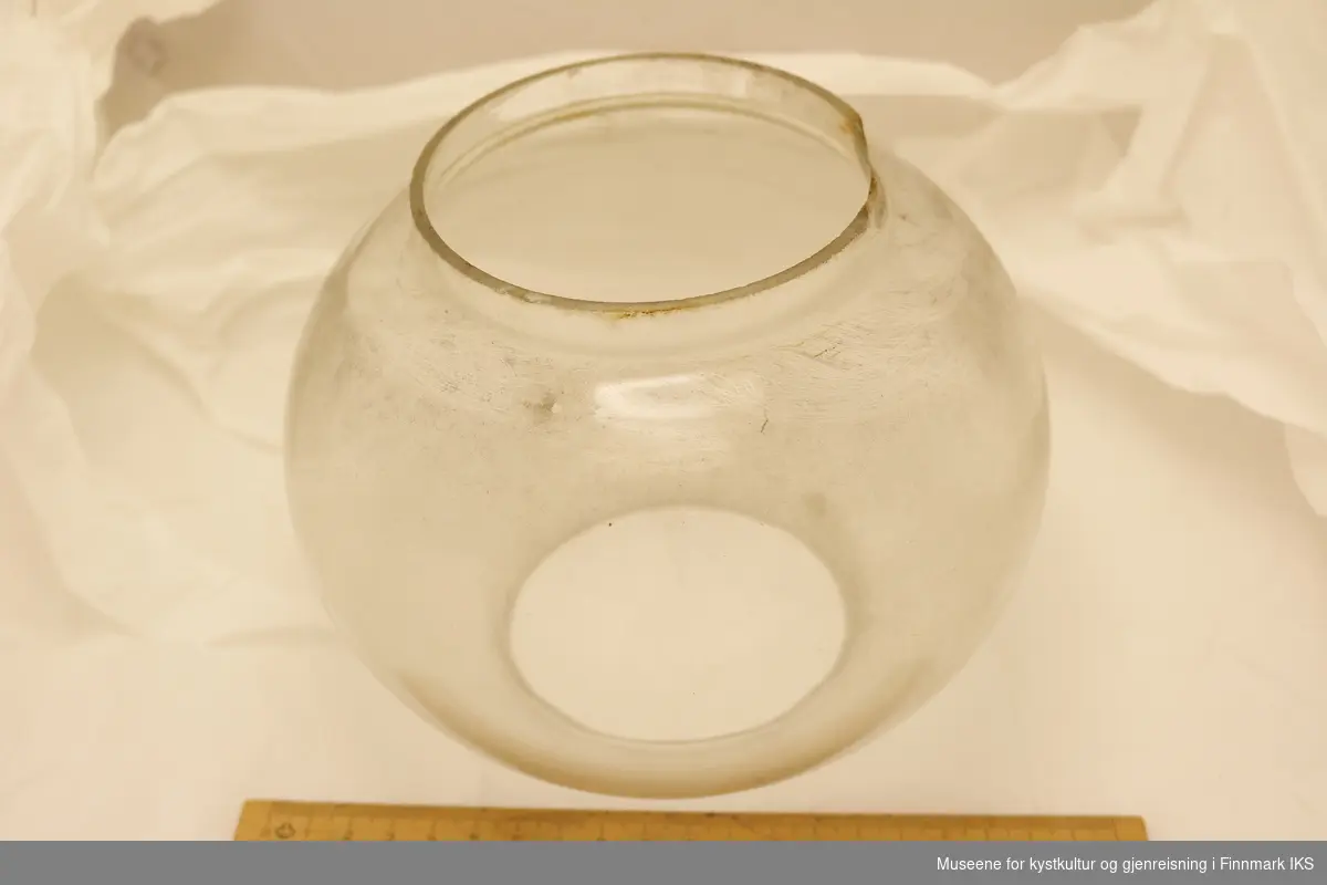 Lampeskjerm i glass med rund form. Åpen i topp og bunn, og har sylinderformet kant langs åpningene. Lampeskjermen er skitten, og har hakk langs kanten i den ene enden.