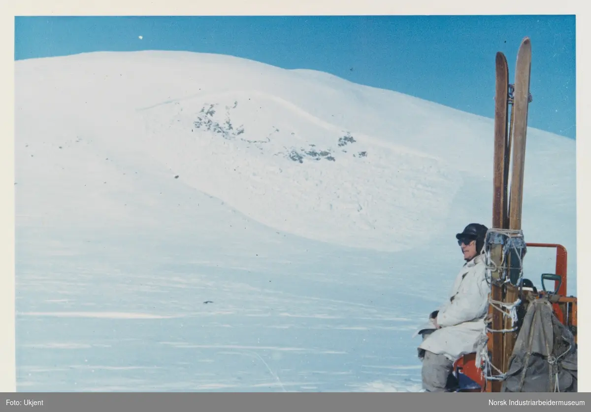 James Coward sitter på snøskuter. Et par ski og ryggsekk er festet på skuteren. Snødekt fjell og blå himmel