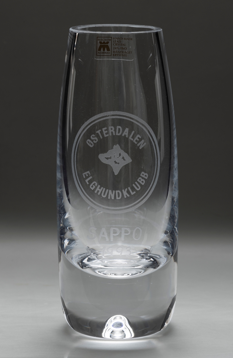 Liten sylinderformet vase i glass, krystall, som er bredest på midten. På den ene siden: innskriften SAPPO / 1993 (med emblemet til Østerdalen Elghundklubb over).