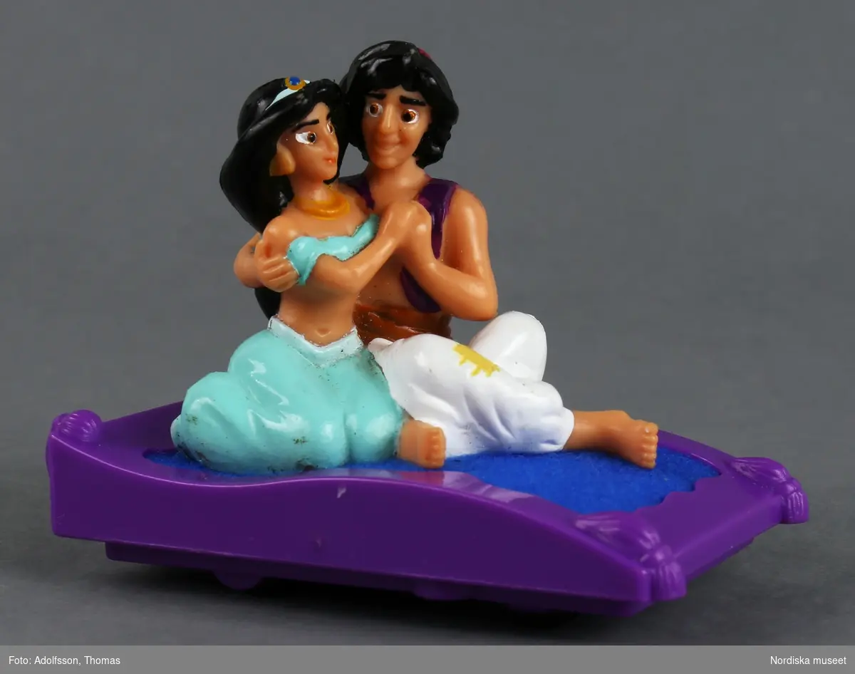Mekanisk leksak med kärleksparet Aladdin och Jasmine på flygande mattan. På undersidan finns fyra hjul, så att leksaken flyger iväg när mekanismen dragits upp.