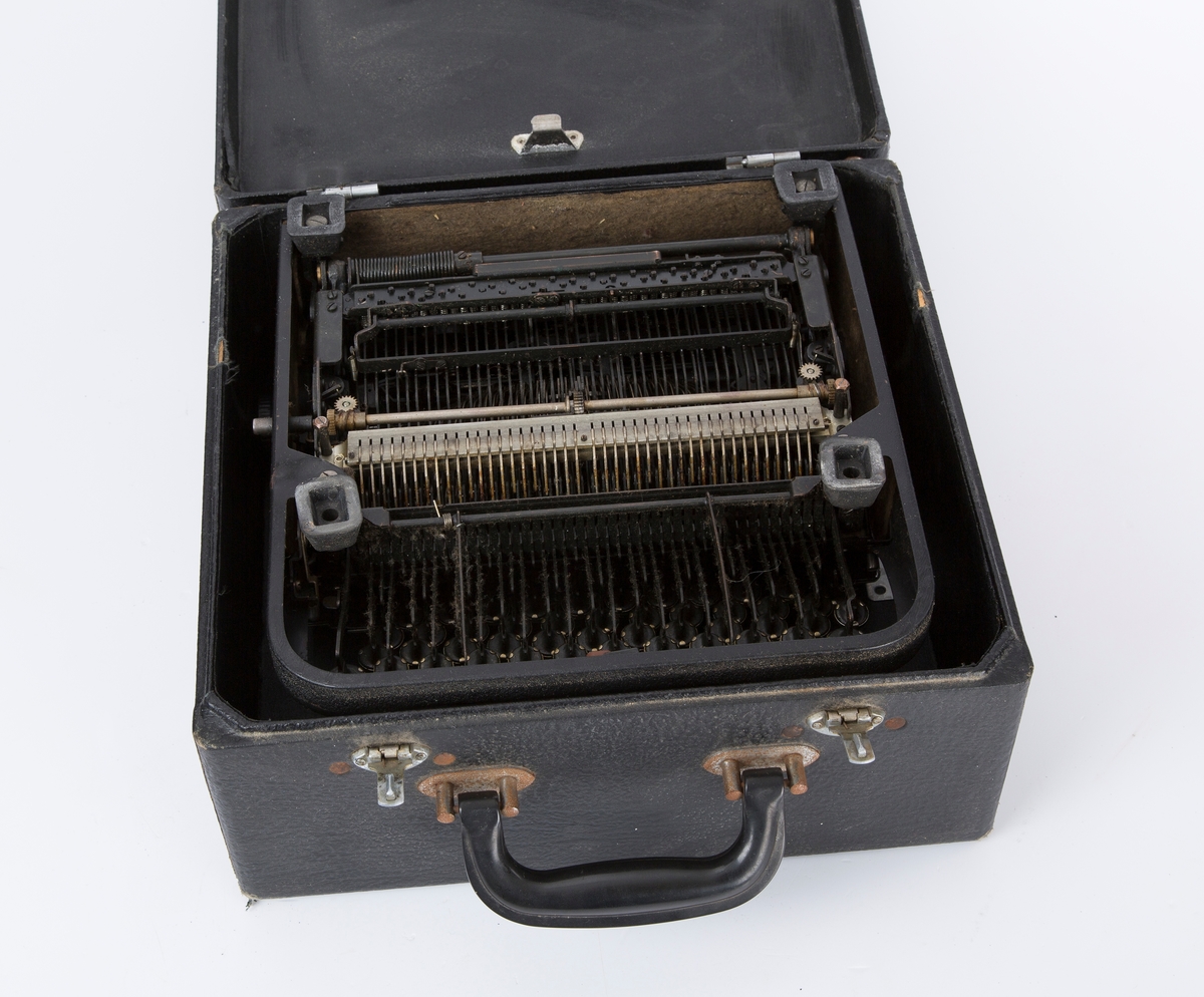 Standard reiseskrivemaskin m/rull og blekkbånd. Maskinen er festet i en kuffert, lokket løftes opp v/bruk. Bærehåndtak. Runde tastaturknotter i fronten m/tall og bokstaver. Fabrikkprodusert.