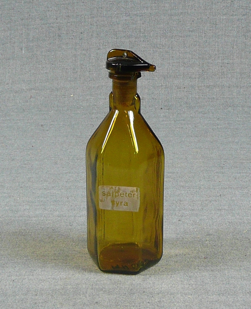 Hexagonal (6-kantig) flaska av brunt glas med texten Salpetersyra. Glaspropp.