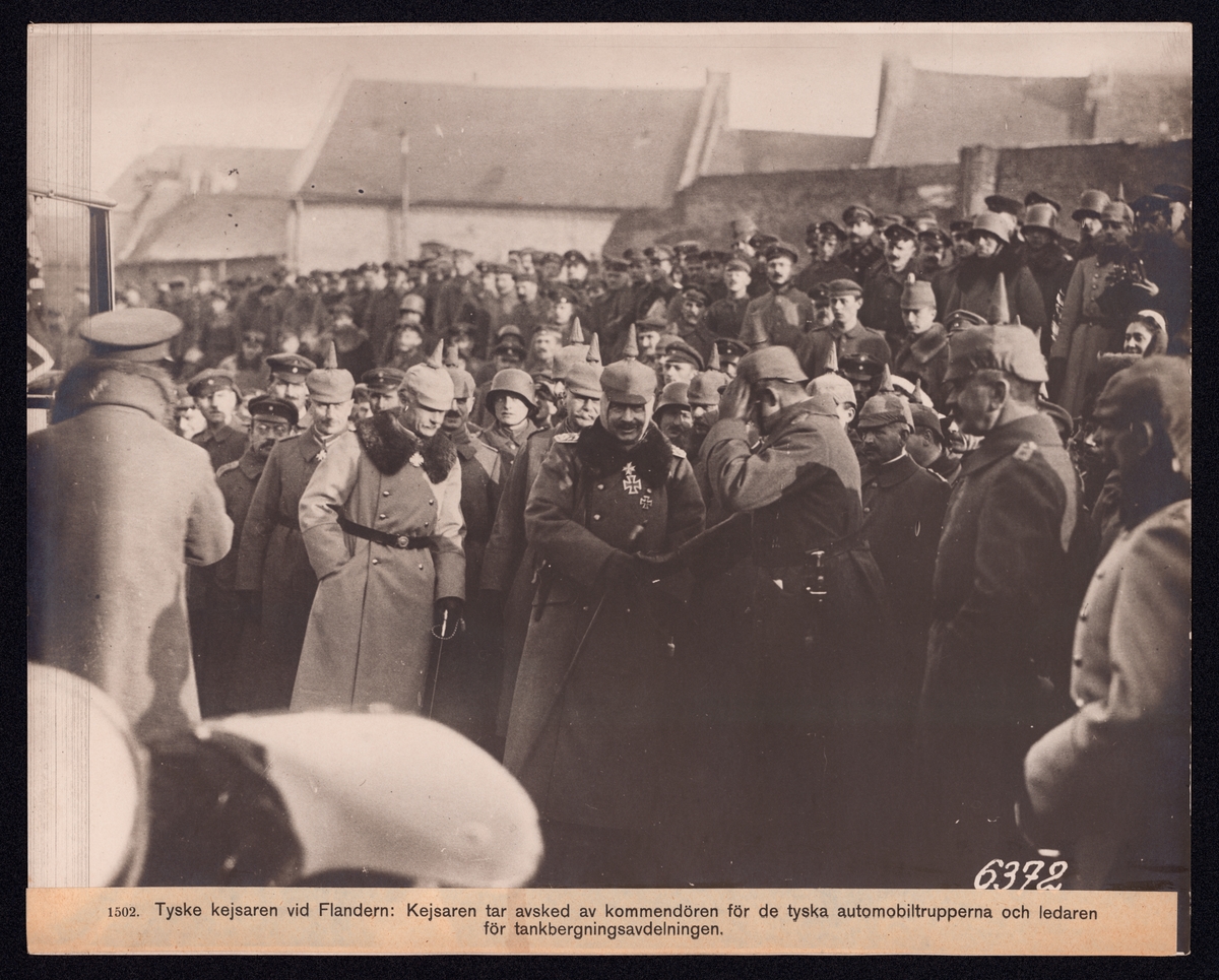 Bilden visar den tyske kejsaren Vilhelm II på ett truppbesök. Han står mitt i en stor åskådarmängd och skakar handen med en officerare. I bakgrunden syns bebyggelse.