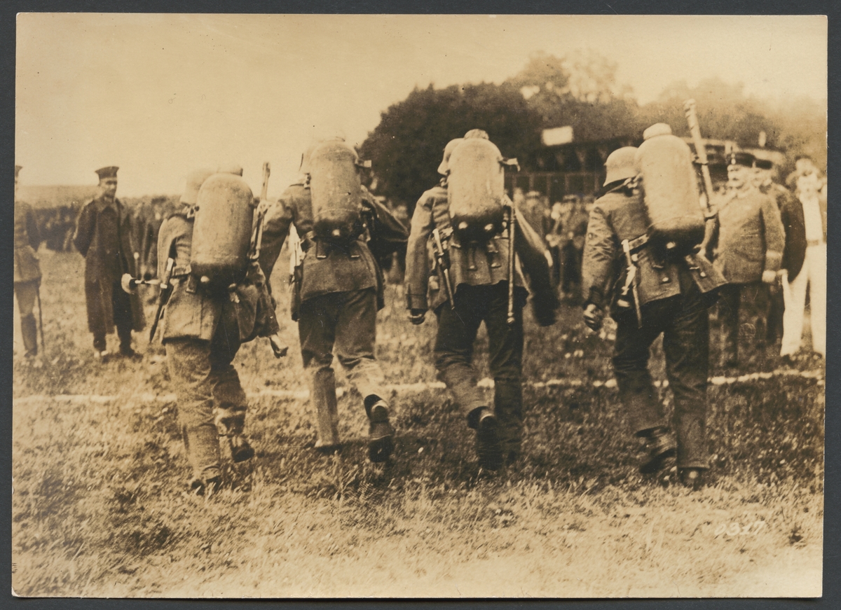 Bilden visar fyra soldater fullt utrustad med uniform, stålhjälm och med eldkastare på ryggen som springer en kapplöpning. Man ser männen bakifrån strax innan mållinjen. I bakgrunden och på sidorna syns åskådare.