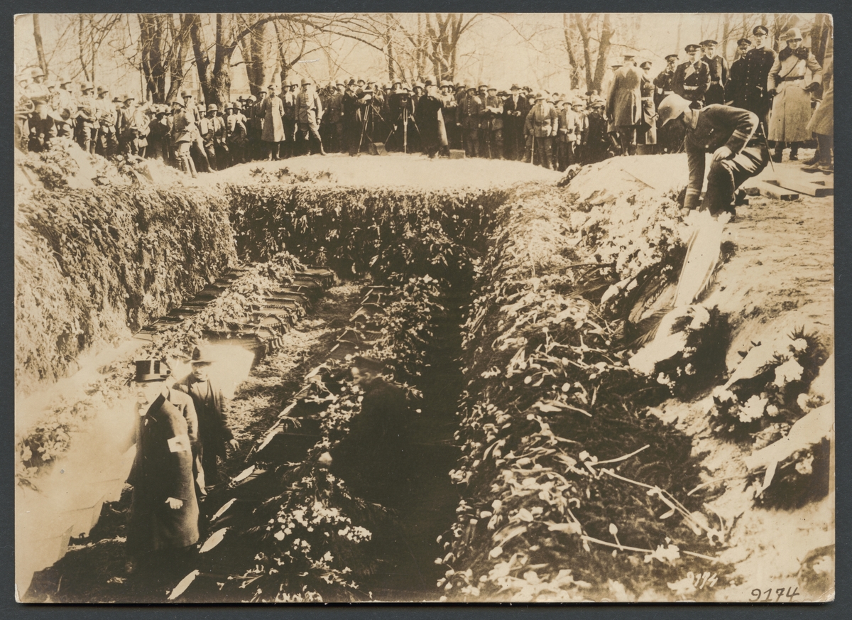 Bilden visar en massgrav med två rad kistor. Graven och kistorna är smyckade med ett hav av blommor. I bakgrunden ser man en större mängd soldater som åskådare och flera fotografer har tagit uppställning. På höger sidan står de högre officerarna.