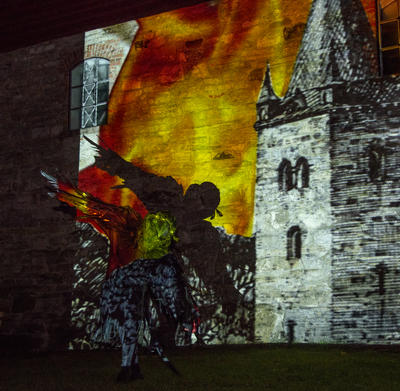 En svart fugl flyr foran en videoprojeksjon av middelalderkatedralen som brenner.