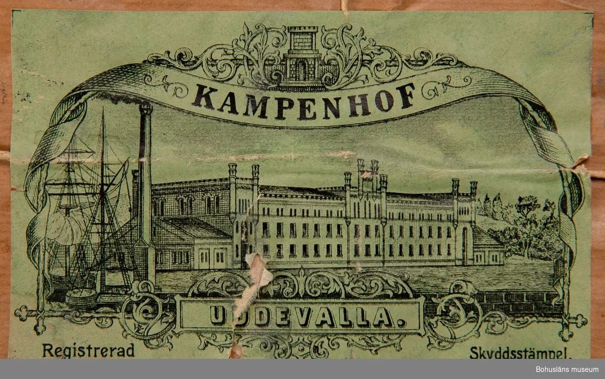 Brunt omslagspapper med firmamärke i grönt papper med bild av Kampenhofs bomullsspinneris fabriksbyggnad.
Text: "Kampenhof, Uddevalla. Registrerad skyddsstämpel."