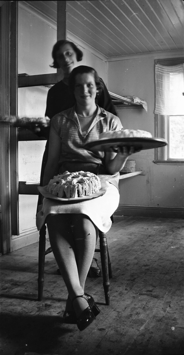 En ung kvinna sitter på en stol i ett kalt utrymme med en tårta i knät och en i handen. Bakom henne står en annan kvinna och håller ett fat med andra bakverk. Bilden kan vara från ålderdomshemmet i Thårstorp.