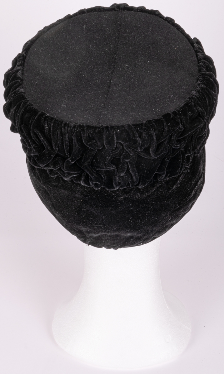 Hatt av svart sammet, utan brätte, nackskydd av sammet. Hattens botten av filt.