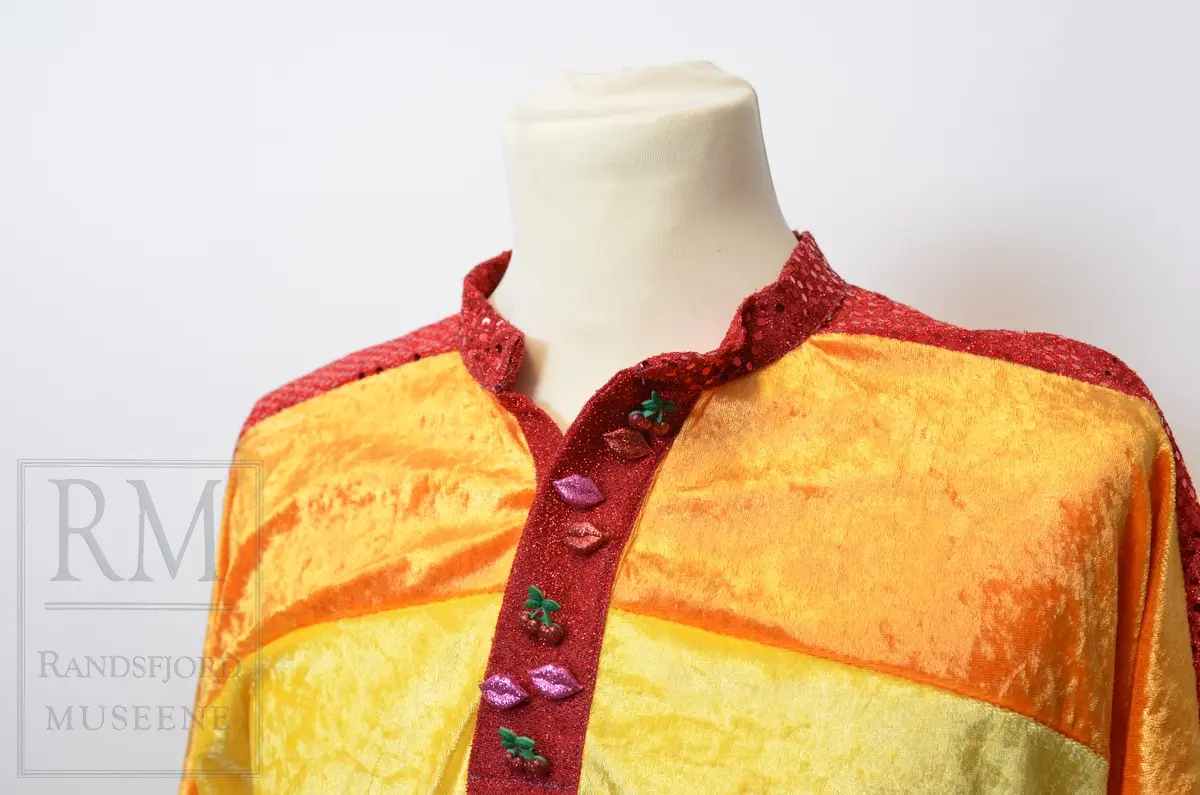 Skjorte i busserull-fasong sydd i regnbuefarget paljettstoff og fløyel. Dekorert med kirsebær- og leppe knapper, samt spensel i ryggen.