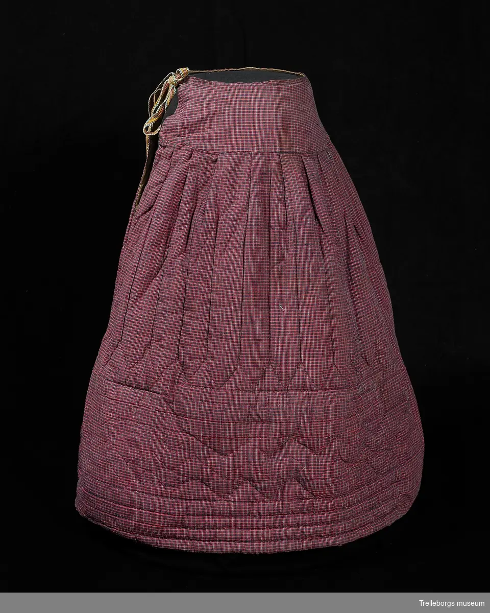 Vadderad handsydd kjol i smårutigt tyg i rött och blått.
Slätt "höftparti" 12 cm, öppet sprund som knytes med ett färgglatt hemvävt(?) band.
Kjolen är fylld med bomullsvadd och häst(?)tagel, fodret är av gråbrunt bomullstyg, dessa tre lager är ihopsydda med olika mönster.