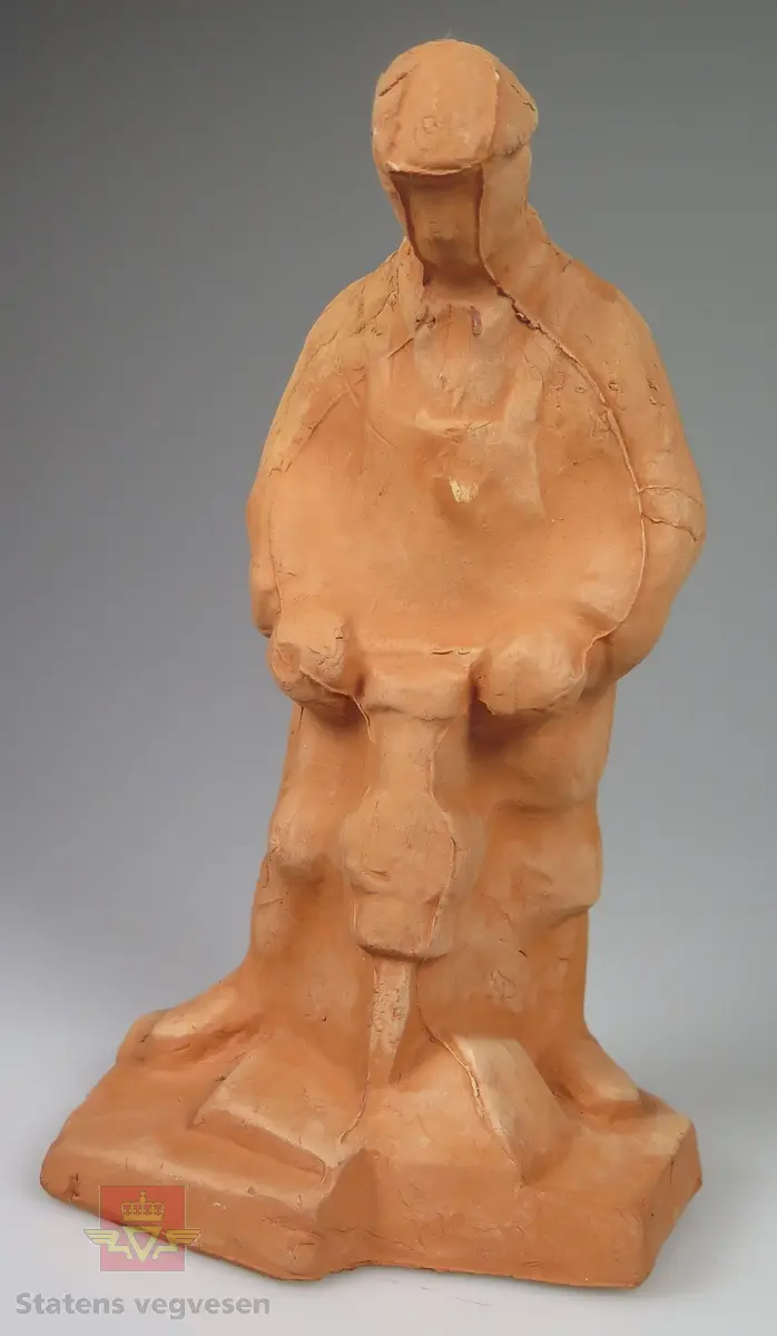 Rødbrun skulptur av en anleggsarbeider med pressluftbor. Anleggsarbeideren har et svakt omriss av klær på seg, og en lue. Detaljnivået er ellers lavt. Under er det grå filt.

Merket: N. AA. 65