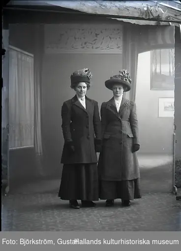 Ateljébild. Två kvinnor i helfigur. Båda bär ytterkläder och hattar. Beställare: Gerda Persson.