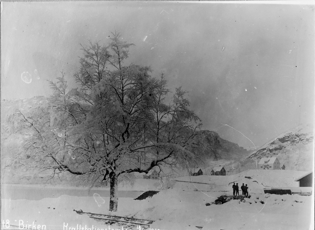 Kraftstasjonstomten med arbeidere vinterstid, Bjørketre i forgrunn, bebyggelse i bakgrunn
Utbygging av Bergen Kraftanlæg i Samnanger