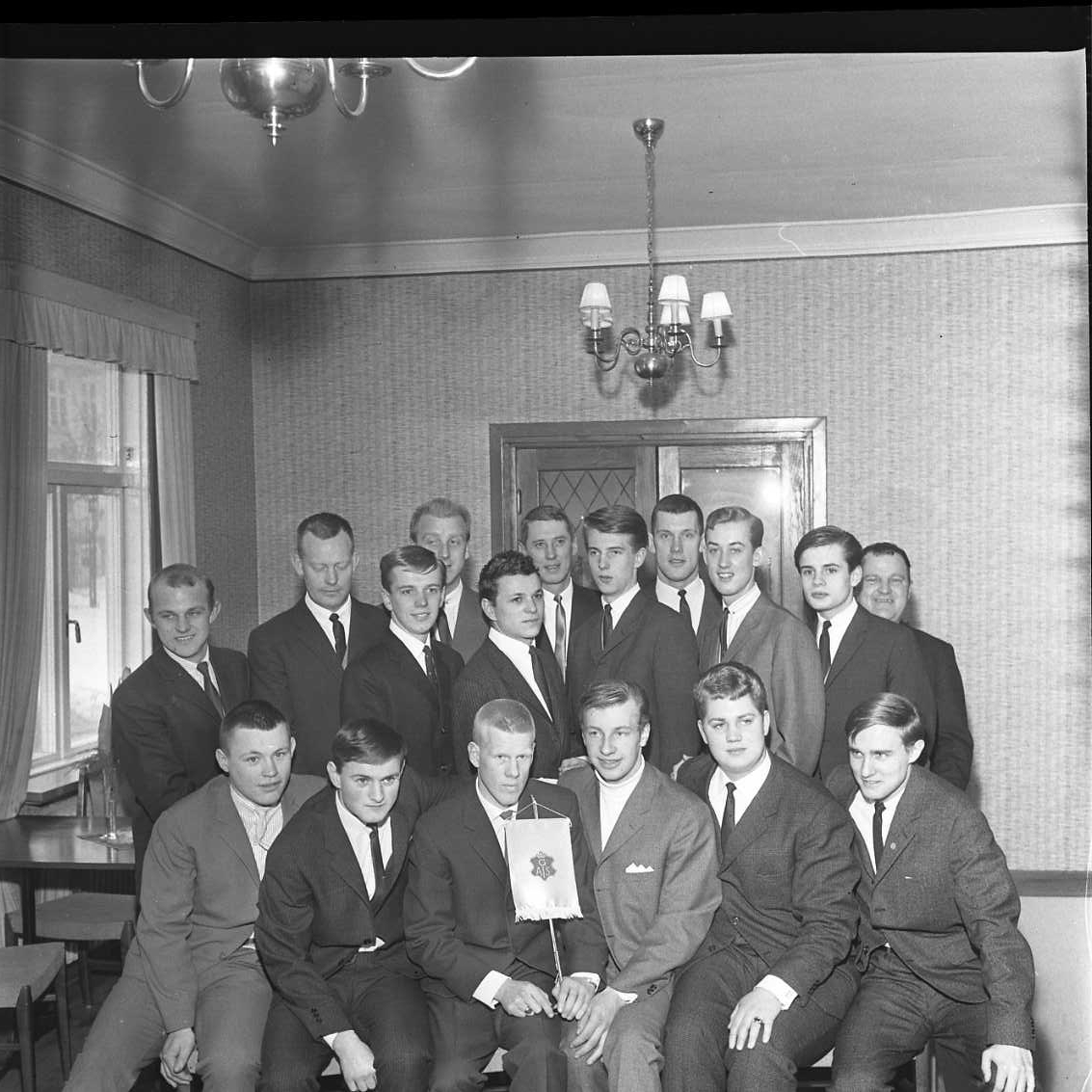 Gruppbild med 17 män, sittande och stående. Två i mitten längst fram håller ett standar med logo för GAIS, det vill säga Gränna AIS. Det är klubbens bandylag årgång 1966. Det är möjligen ett fotografi i samband med årsmöte för Gränna AIS.