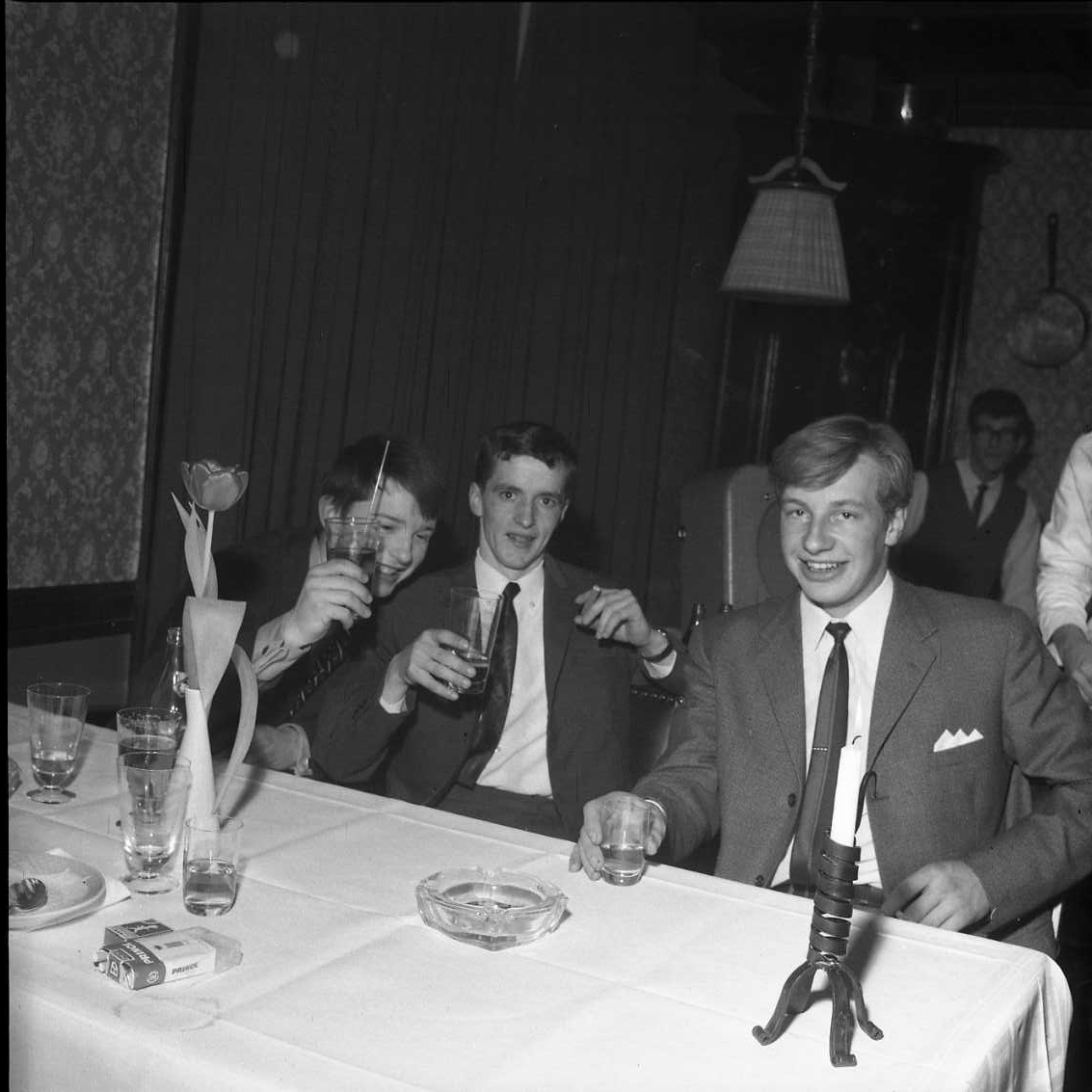Tre unga och glada män i kostym och slips sitter vid ett bord med glas i händerna. Mannen till vänster lyfter sitt glas som en skål-gest mot fotografen. På bordet står det flera tomma glas, askkopp, cigaretter och några använda assietter. Fotografi i samband med årsmöte för Gränna AIS.