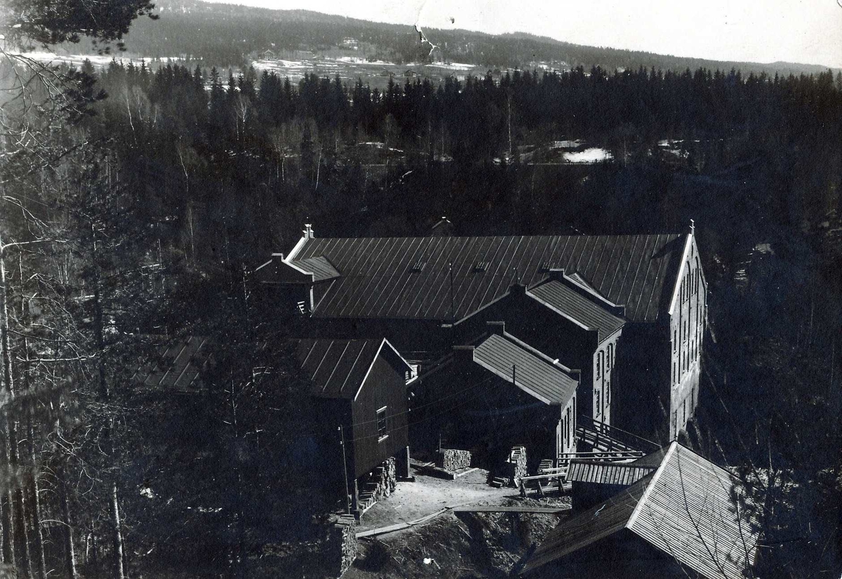 Industrianlegg med bygninger av tegl og tre på A/S Kistefos Træsliberi. Skog, dyrket mark og jernbanetrasé i bakgrunnen.