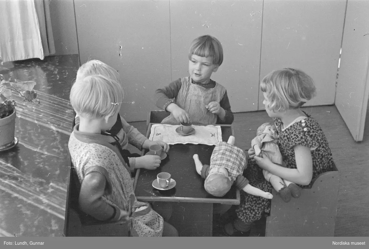 Motiv: H.S.B.s lekstuga på Skeppargat. (gärdet) (Skeppargatan) (HSB);
Ett barn leker i en leksaksbåt, barn leker med dockservis och dockor, två barn har kronor av papper, två barn kramas.