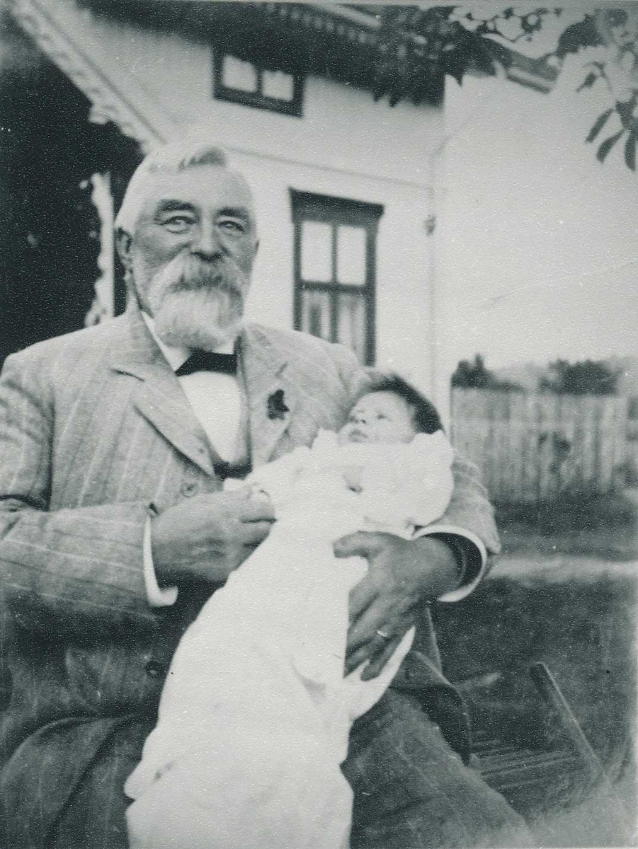 Eldre mann i dress sitter utendørs med en baby i armene. 
