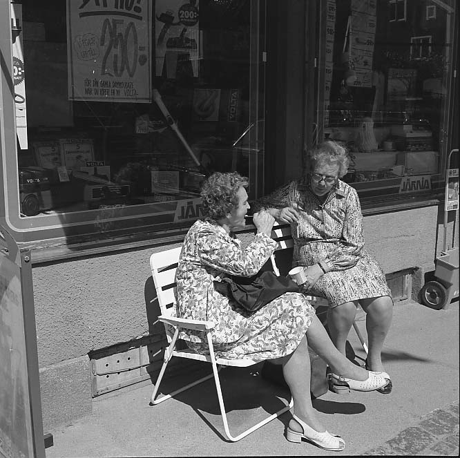 Två kvinnor i mönstrade klänningar sitter i varsin stol utanför skyltfönstret till Palm Järn på Brahegatan i Gränna. Kvinnan till vänster äter möjligen glass ur en bägare. De sitter delvis i skugga.