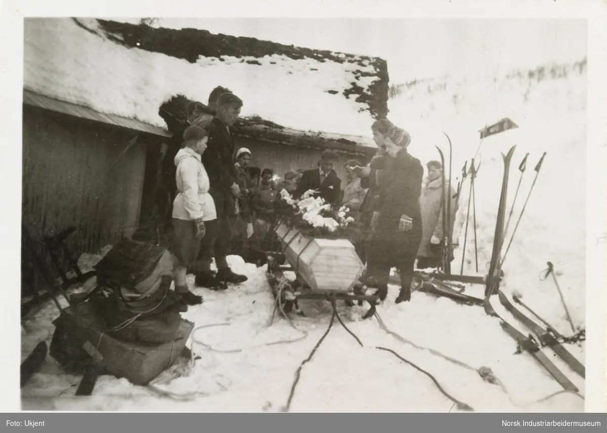 Begravelsen til Bergit Larsen. Kiste liggende på slede i snøen utenfor hus. Rundt står forsamling med mennesker, ski og staver