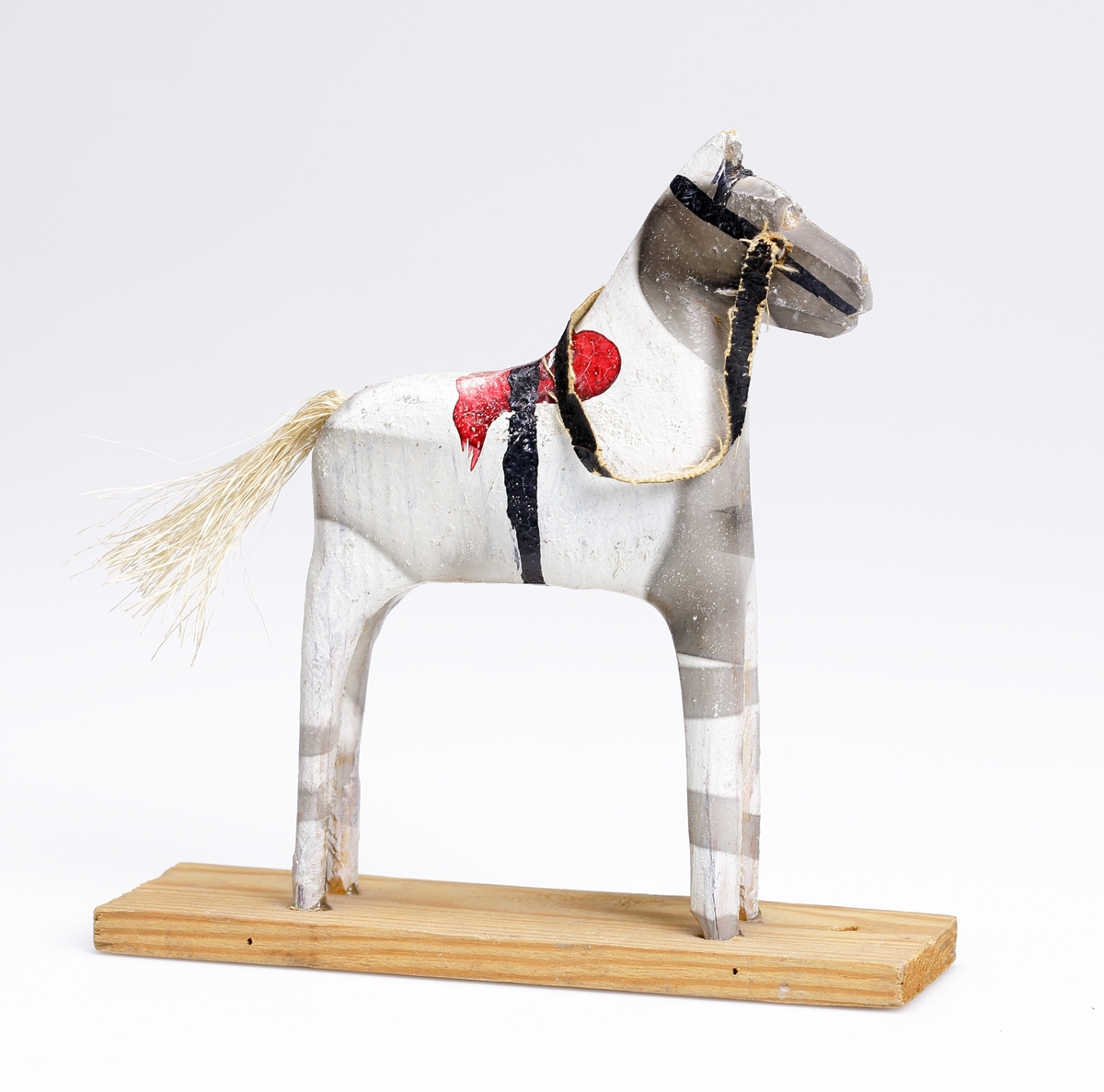 En snidad leksakshäst av trä och med en svans av tagel. Målad vit, grå, röd och svart.
