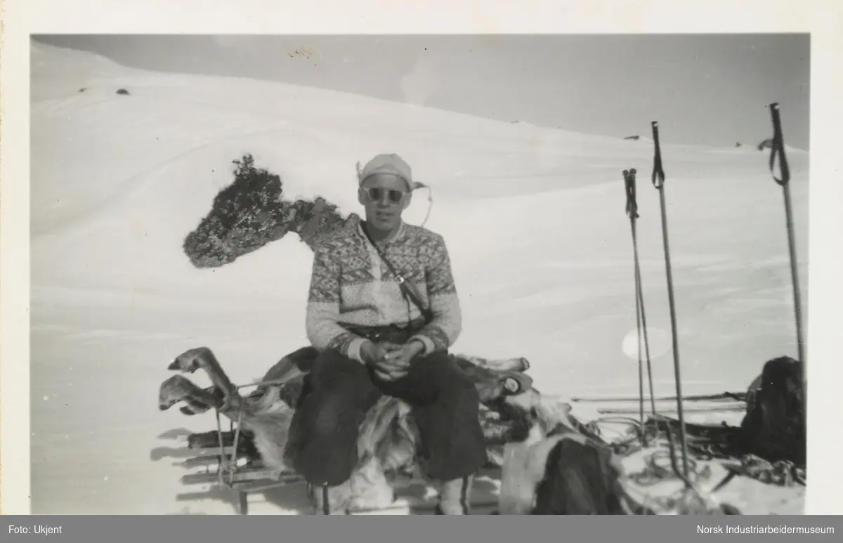 Olav O. Vågen med lusekofte og solbriller sitter på et felt reinsdyr som ligger på en slede i fjellet. Ved siden ligger ski, skistaver og ryggsekk
